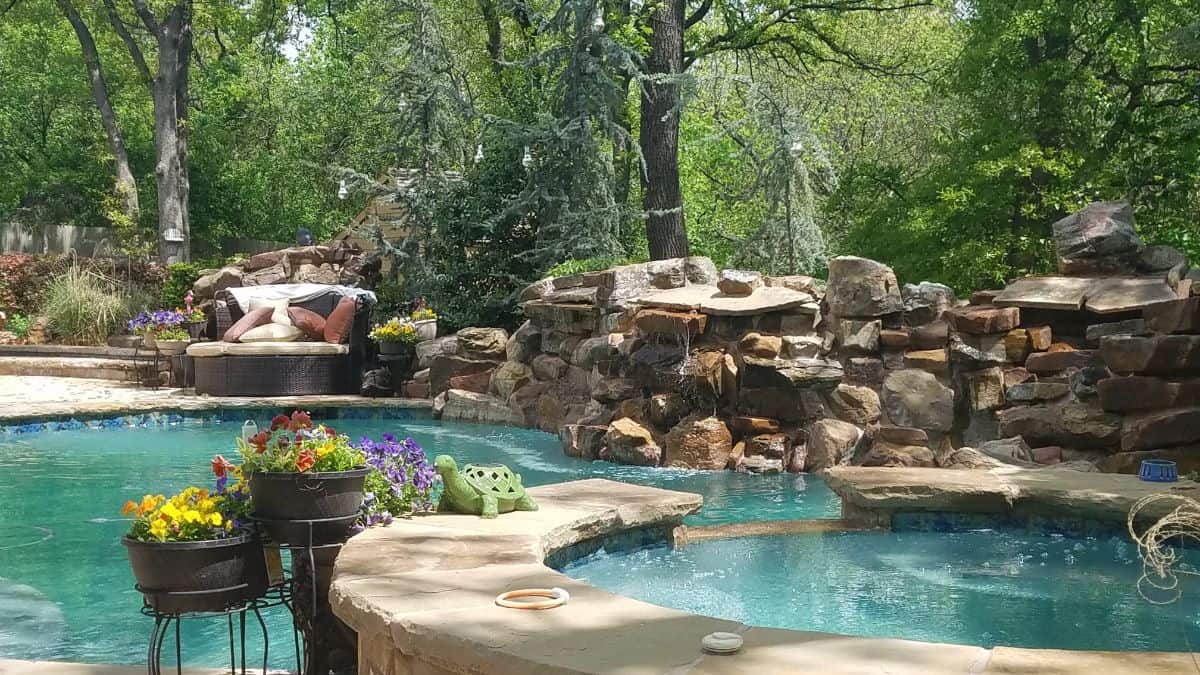 Rear view of a 90 ft raised freeform pool with rock waterfall and grotto, surrounded by landscaping and a love seat.